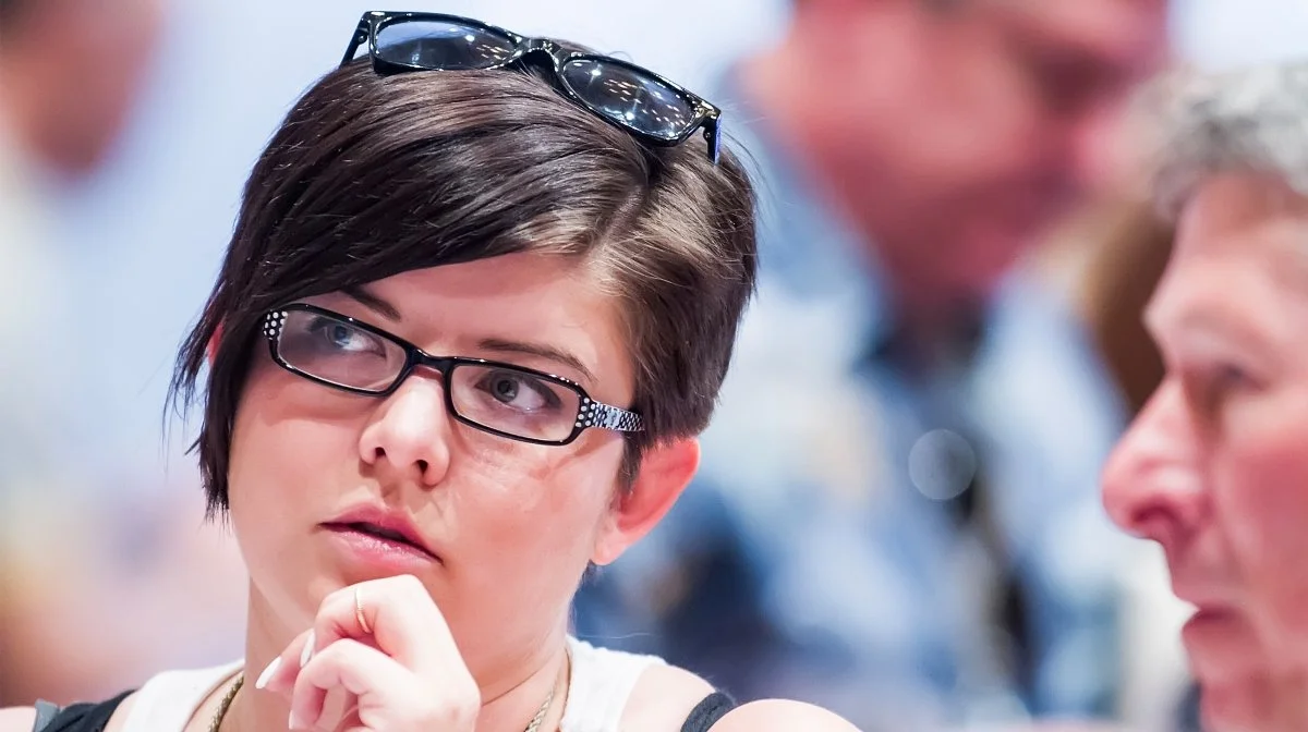 A woman with short dark hair and glasses, thinking, with a group of people blurred in the background.