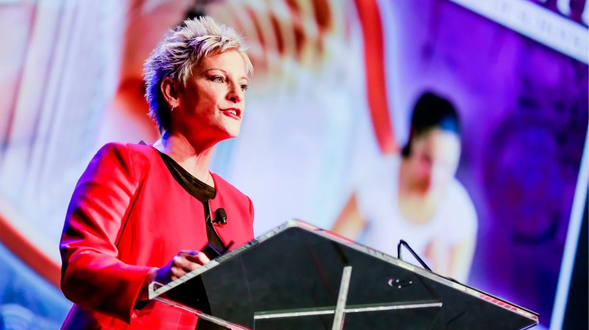 A woman with short blonde hair in a red blazer giving a presentation at a conference.