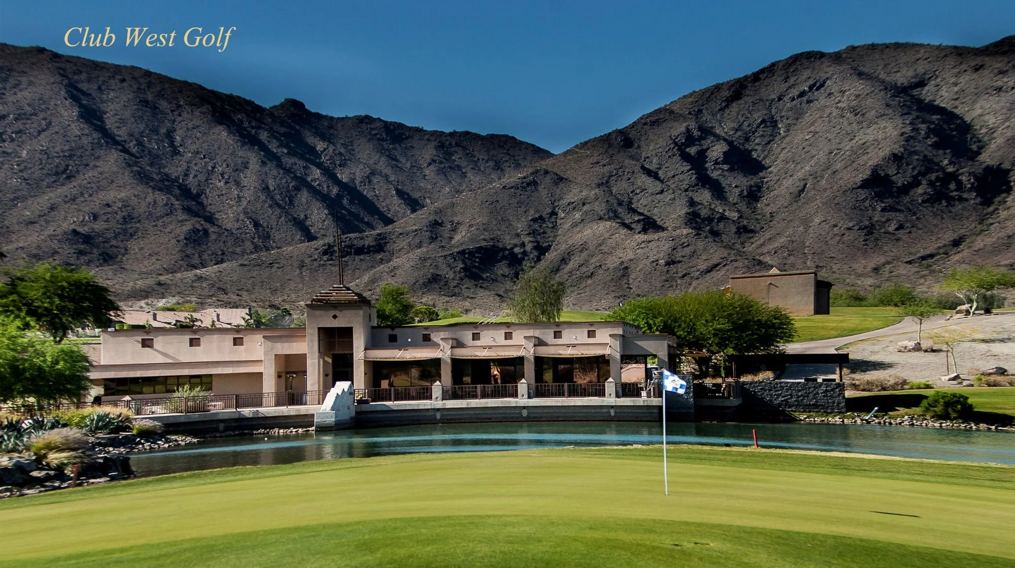 A view of a golf course at Club West Golf with a mountain range in the background. There is a building near the water hazard with a flagstick on the green in the foreground. The sky is clear and blue.