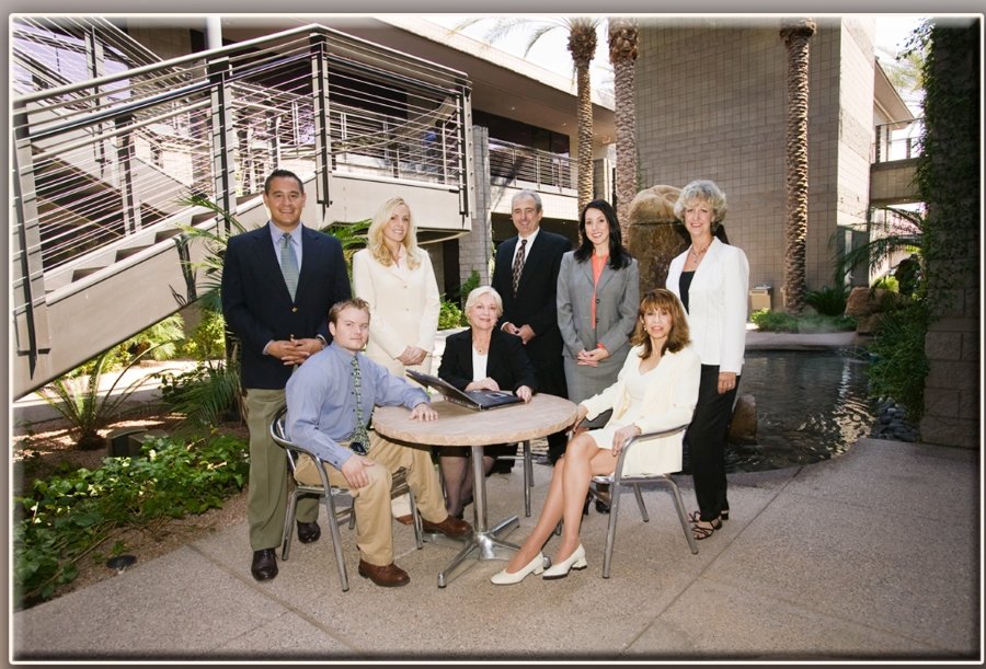 Group of nine professionally dressed people gathered around a table outdoors in a modern courtyard with palm trees and water feature.