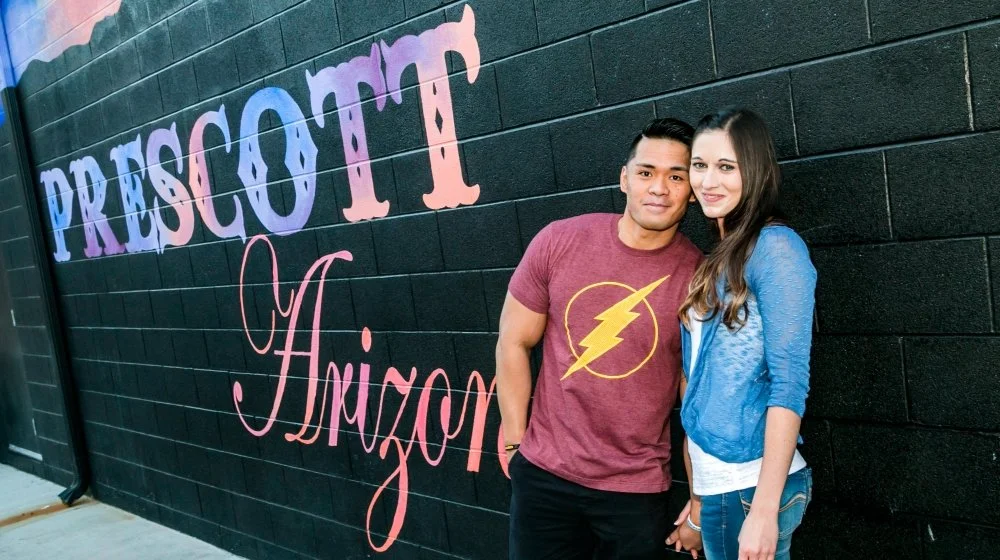 A young man and woman standing close together in front of a black brick wall with colorful writing that says 'PRESCOTT ANIZON.'