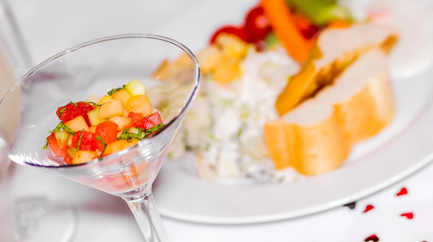 A cocktail glass filled with a colorful vegetable salad, with a blurred plate of sliced vegetables and a salad with dressing in the background.