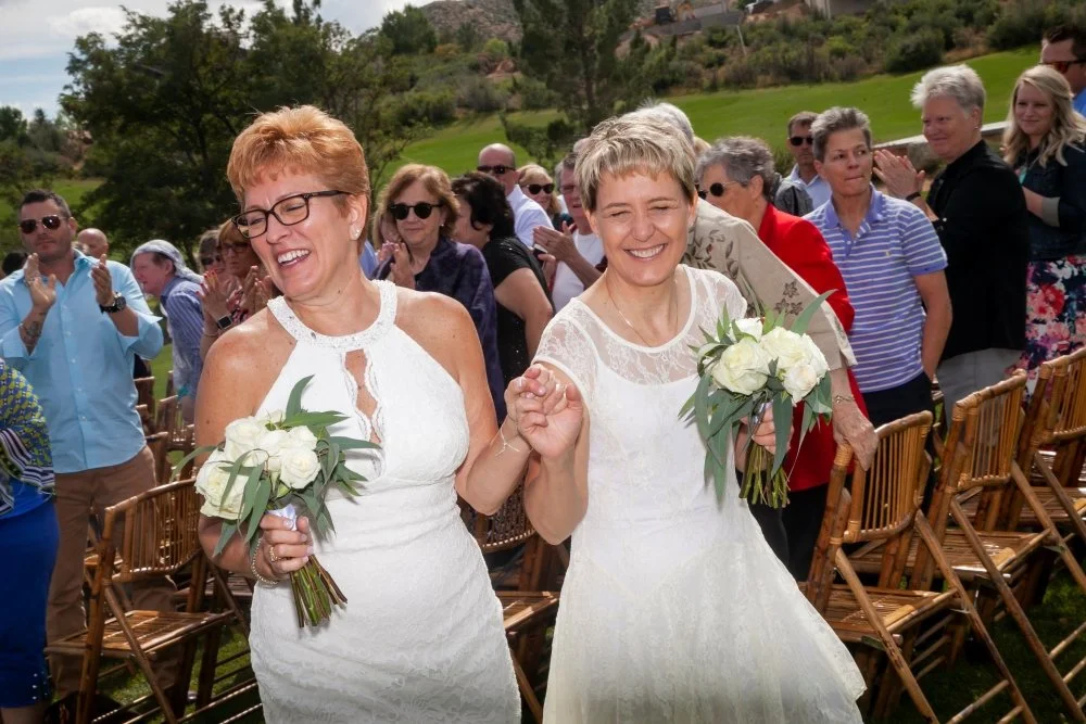 Two women in white dresses holding bouquets, smiling and holding hands at an outdoor wedding ceremony on a grassy hill, with guests clapping in the background.