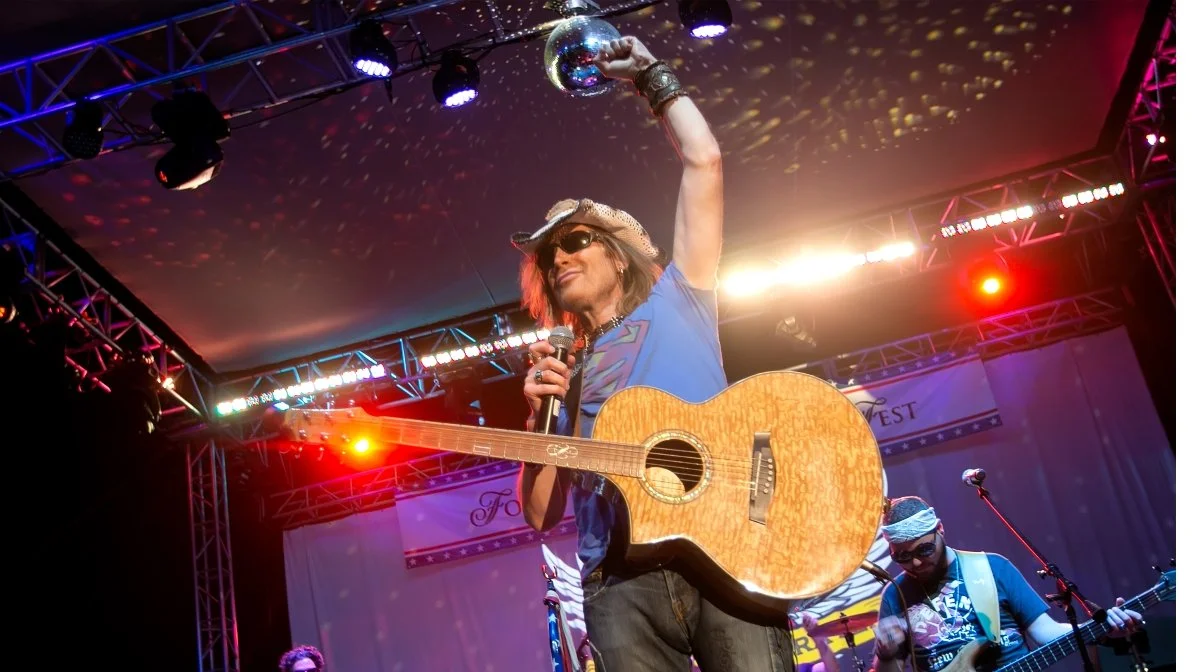 Musician wearing sunglasses, cowboy hat, and blue t-shirt on stage, holding an acoustic guitar in one hand and a microphone in the other, with a disco ball overhead and stage lights.