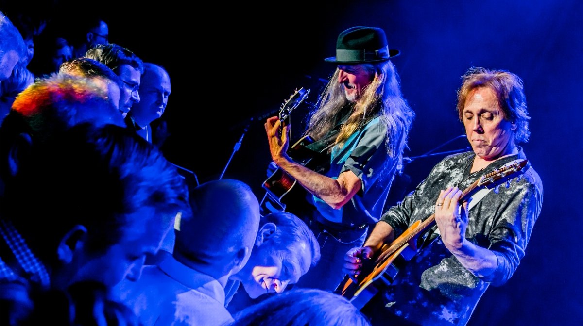 Two musicians playing acoustic guitars on stage with an audience watching, with dramatic blue and purple lighting.