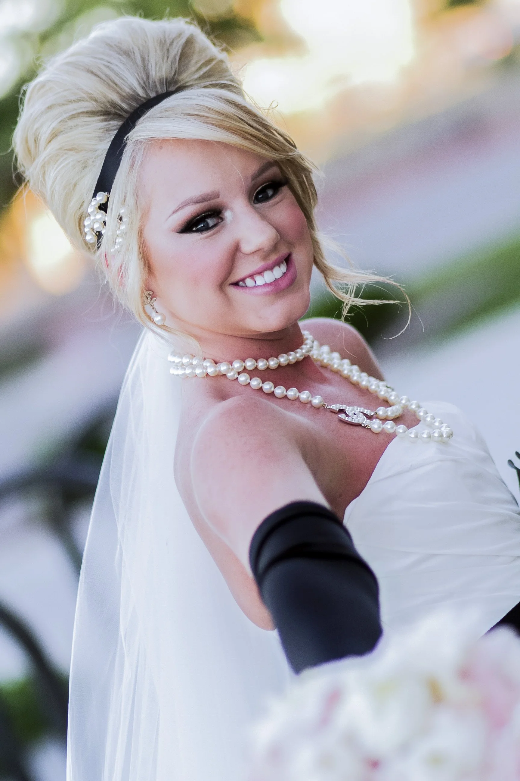 A smiling woman wearing a wedding dress and pearl jewelry taking a selfie outdoors.