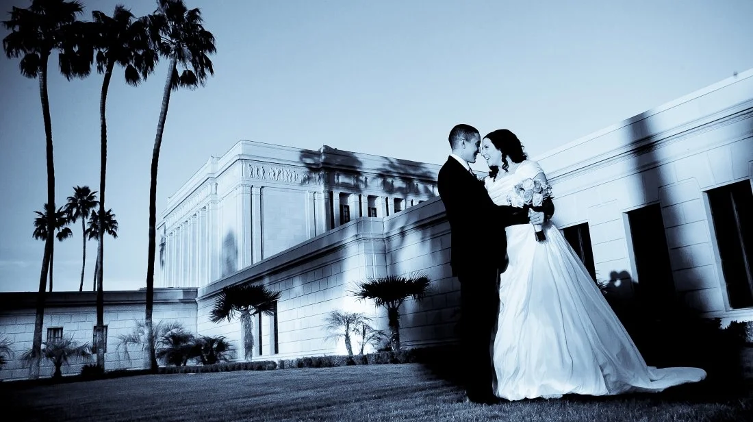 A bride and groom in wedding attire sharing an intimate moment outdoors at night, with palm trees and a large building in the background.
