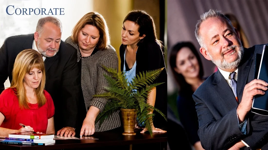 Group of professionals, including men and women, gathered around a table with a woman holding a pen and looking at documents, with a potted fern plant in the center, in an office setting. To the right, a man in a suit is holding a tablet and smiling,