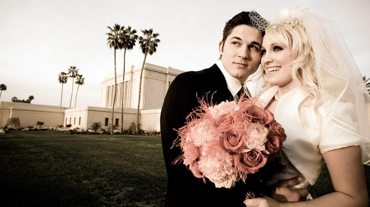 A newlywed couple standing outdoors on a lawn, with palm trees and a large building in the background. The groom is wearing a black tuxedo, holding a bouquet of pink and peach flowers, and the bride is in a white wedding dress with a veil and decorat