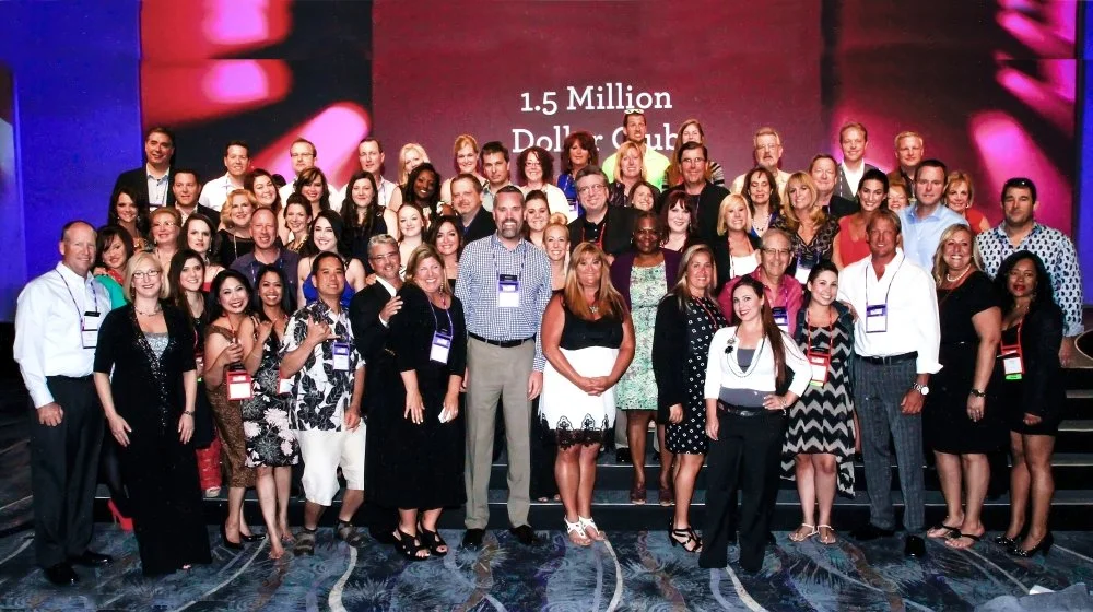 Group of smiling people at a conference or event, with a large screen behind them displaying '1.5 Million Dollar Club'.