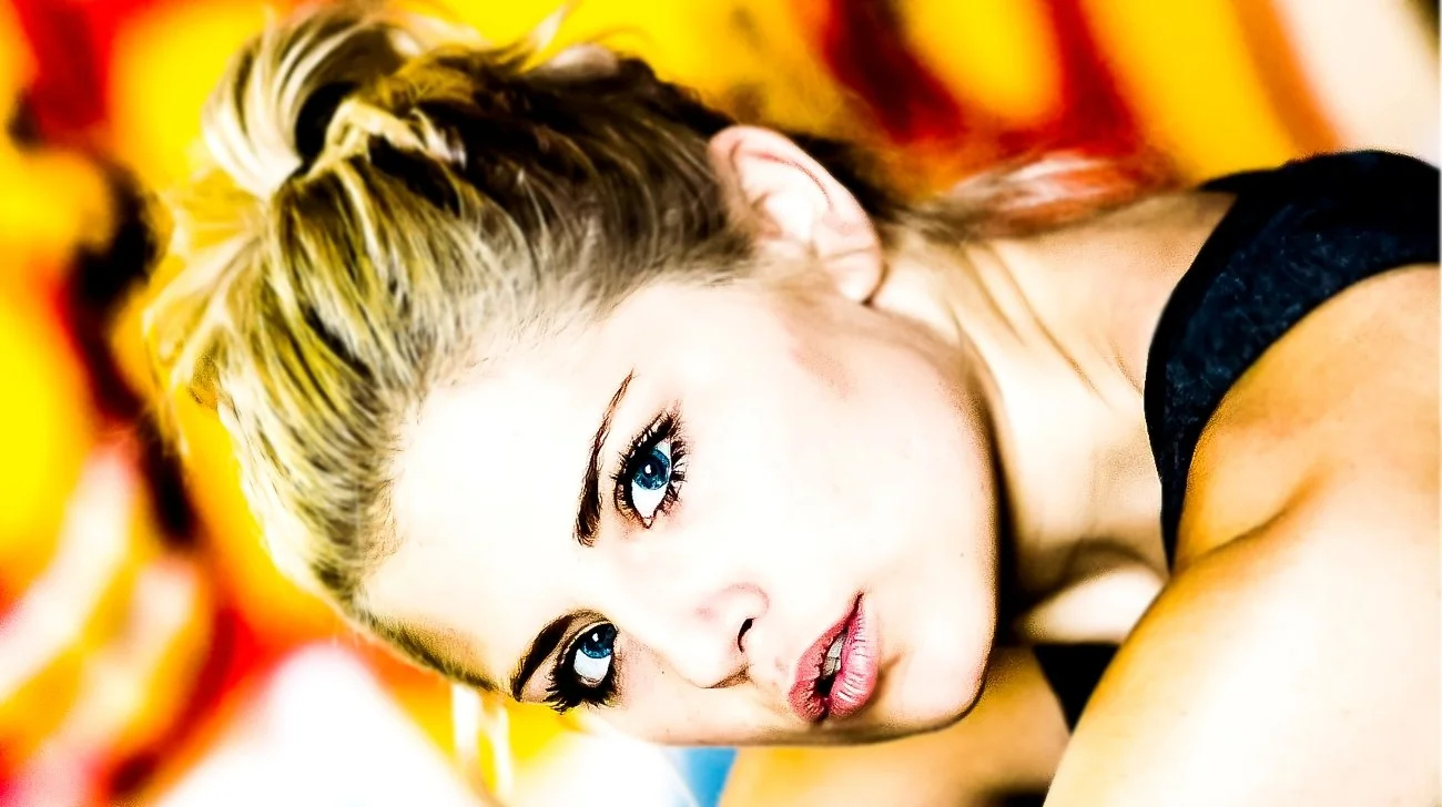 Close-up of a woman with blonde hair and blue eyes lying on a colorful surface, looking at the camera.