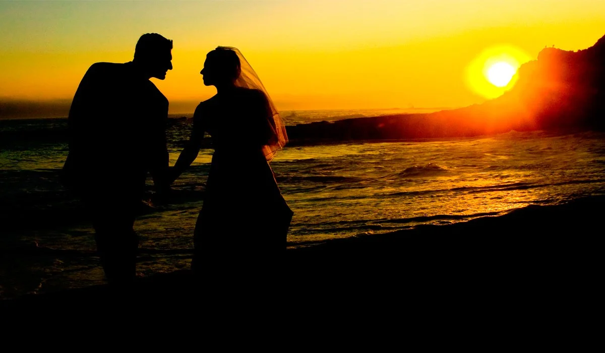 Silhouette of a couple holding hands on the beach at sunset, with the ocean and sun in the background.