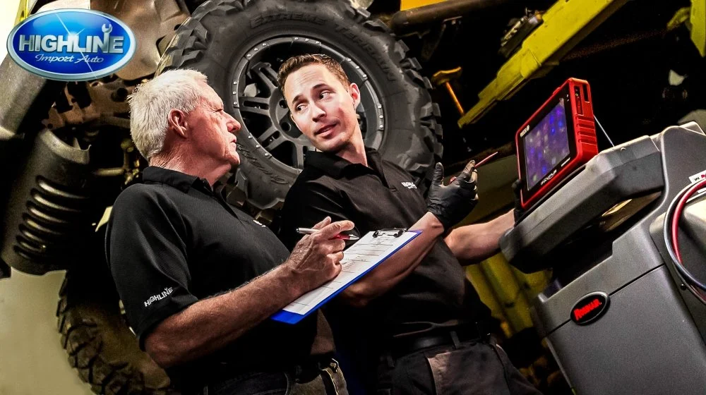Two men working in an auto repair shop, discussing vehicle diagnostics using a digital monitor. The older man holds a clipboard, and the younger man points at the screen. Large tire and vehicle parts are visible in the background with a Highline Impo