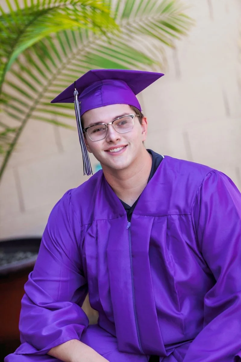 Young man in purple graduation gown and cap smiling, sitting outdoors with green palm leaves and beige wall in background.