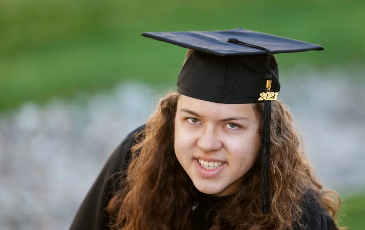 Young woman in graduation cap with '2021' charm, outdoors, smiling.
