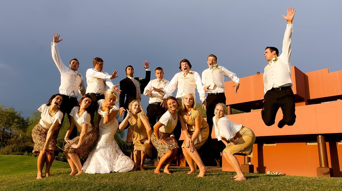 Group of diverse people at a wedding, some in traditional attire, jumping and posing on grass with a modern building and cloudy sky in the background.