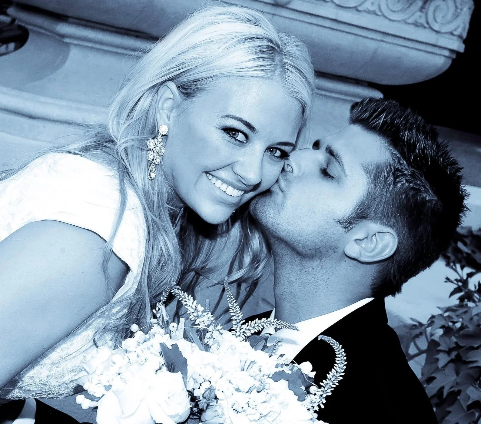 A smiling blonde woman in a wedding dress holding a bouquet of flowers and a man in a tuxedo kissing her on the cheek.
