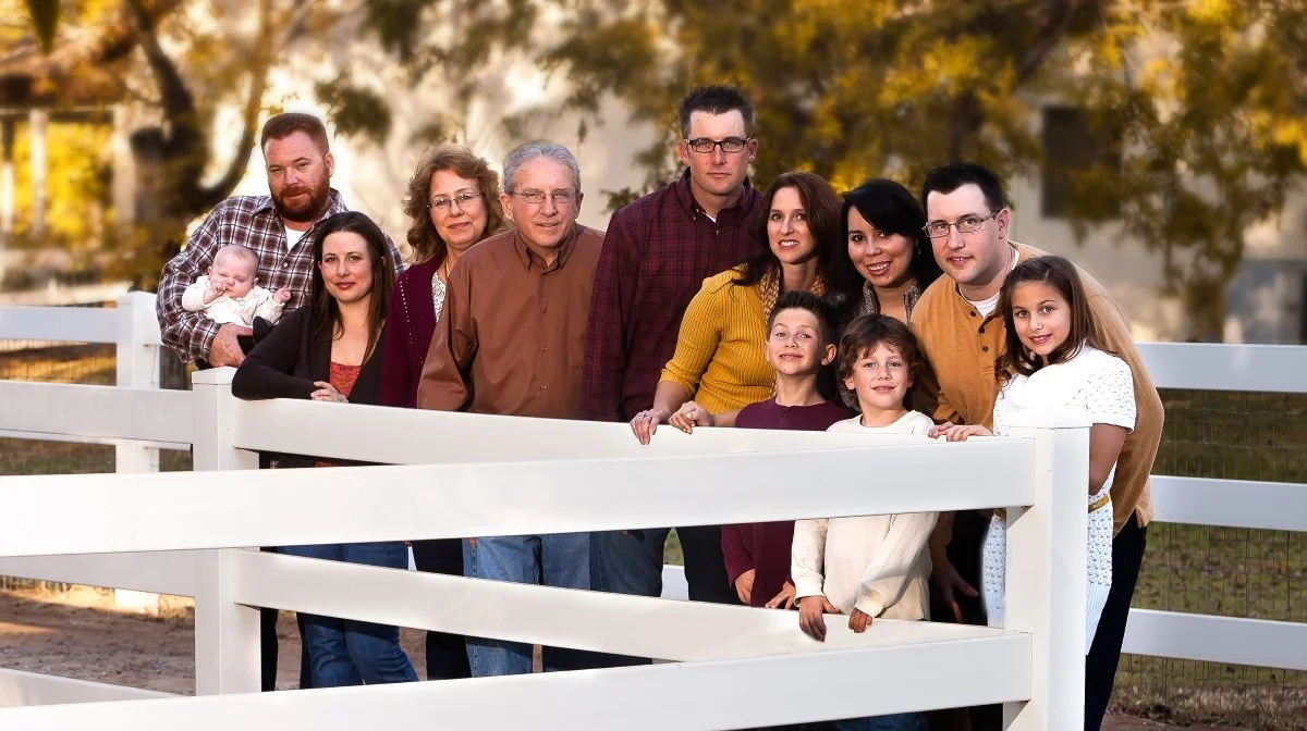 Family photo outdoors with adults and children standing behind a white fence, autumn trees in the background.