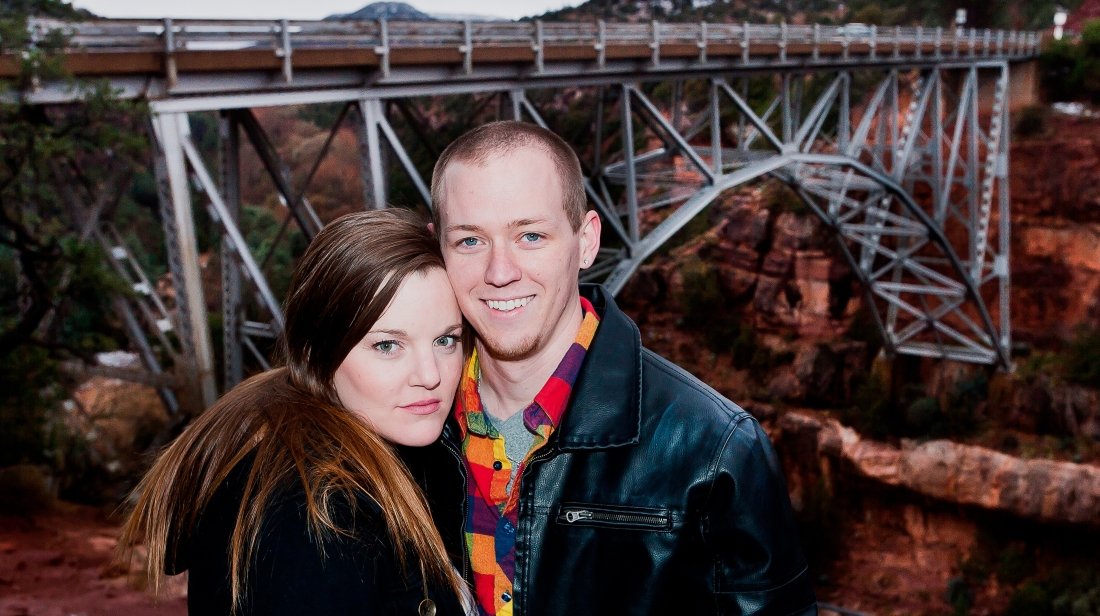 A young couple standing close together outdoors near a bridge, with the man smiling and the woman looking serious.