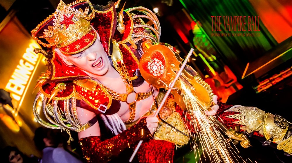 Person dressed in elaborate, colorful costume with ornate headdress, engaging in performance with sparks flying, at the Vampire Ball event.