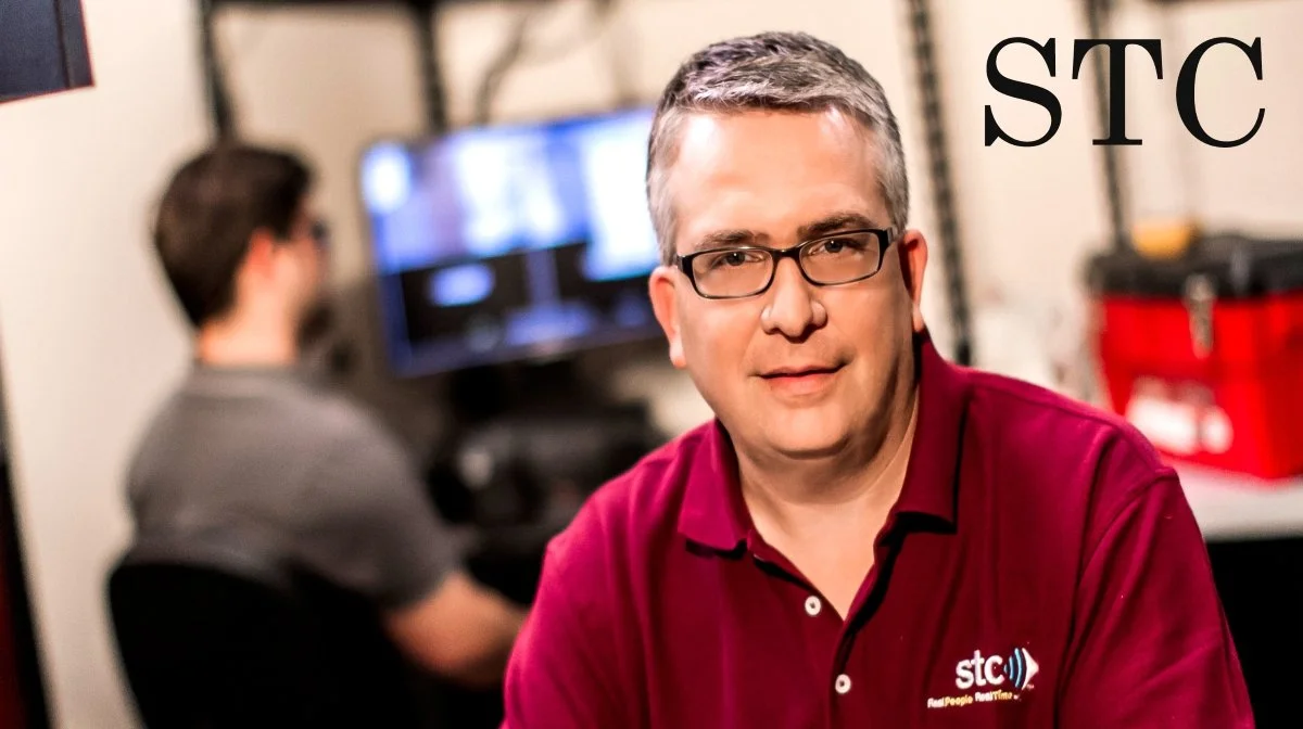 A man with glasses and short gray hair wearing a maroon polo shirt with the STC logo, sitting in a room with blurred people and equipment in the background, including a computer monitor and a red toolbox.