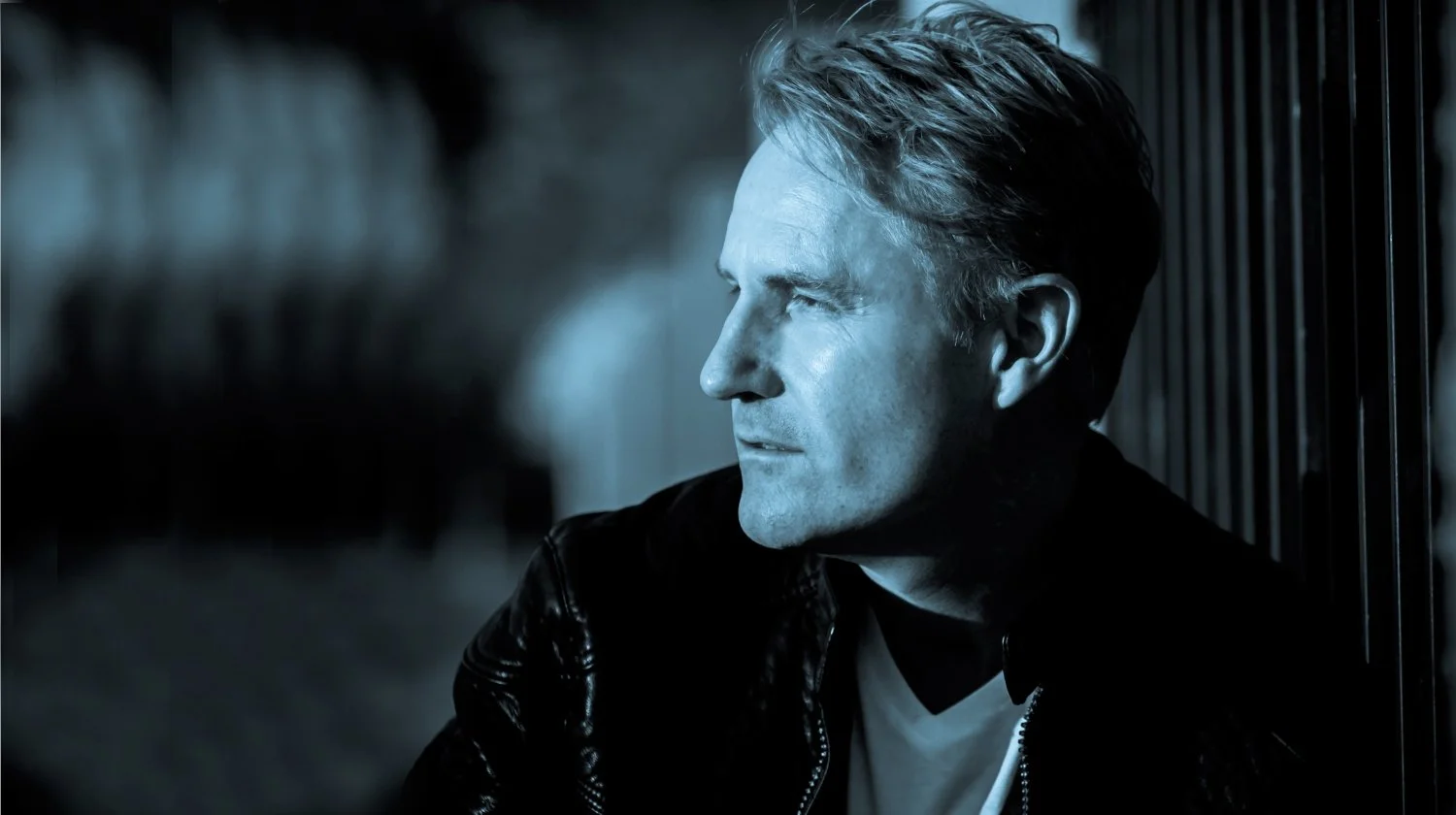 A man with blond hair, wearing a black leather jacket and white shirt, sitting outdoors at night near a metal fence, looking to the side with a serious expression.