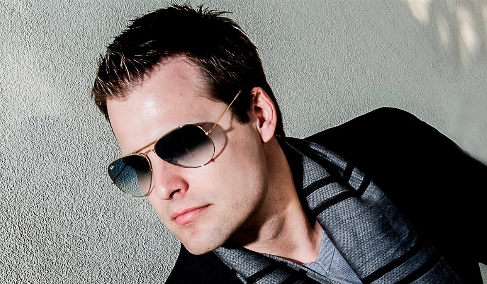 A man wearing sunglasses, a dark jacket, and a striped shirt in front of a textured wall.