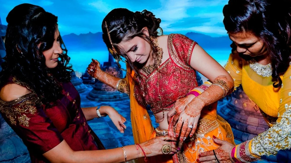 Three women in traditional South Asian attire, focusing on henna application on a bride's hands, during a cultural celebration.