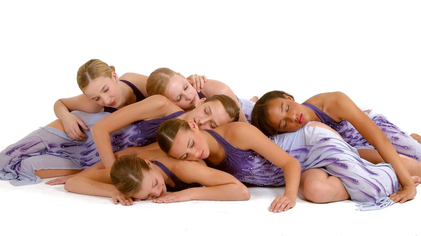Six women wearing matching lilac and purple dresses, lying on the floor and intertwined, appearing to be posing in a choreographed dance or artistic formation.