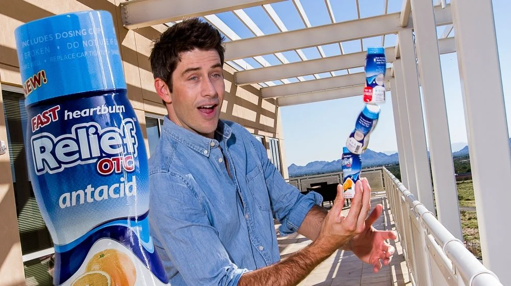 A man balancing several cans of Relief Od pain relief antacid medicine on his hand on a balcony with a desert landscape in the background.