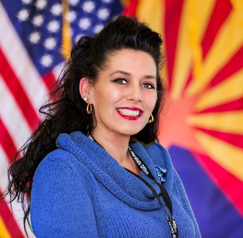 A woman with dark, curly hair and red lipstick, wearing a blue blazer and earrings, smiling in front of an American flag and another flag with red and yellow rays.