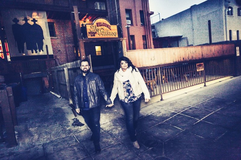A man and woman holding hands walking outside a bar at night, with a sign that reads "The Palace" and a silhouette artwork of three figures in the background.