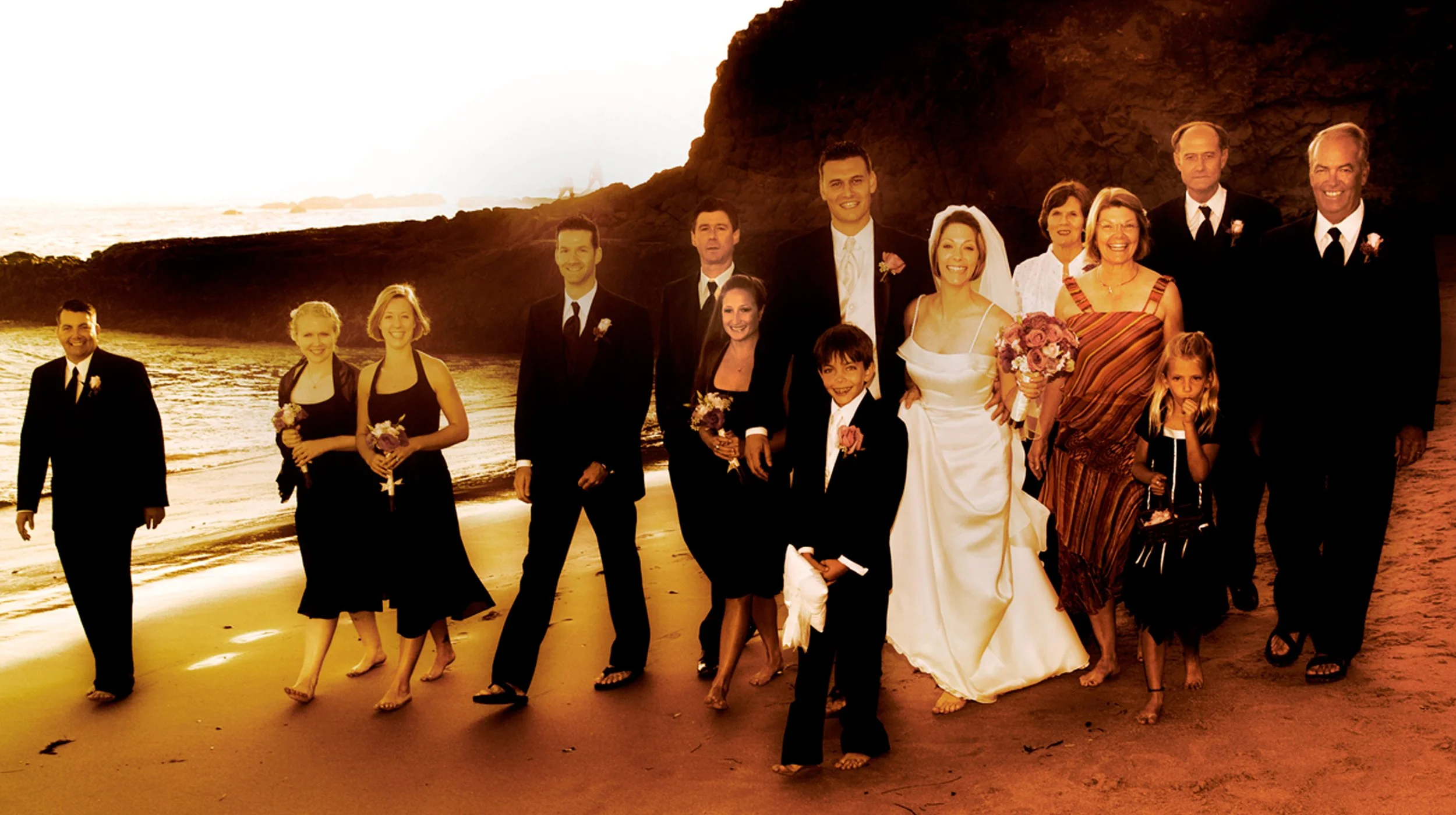 A large group of people dressed in formal attire, including a bride in a white gown and guests in suits and dresses, walking along a beach during sunset, with cliffs and the ocean in the background.