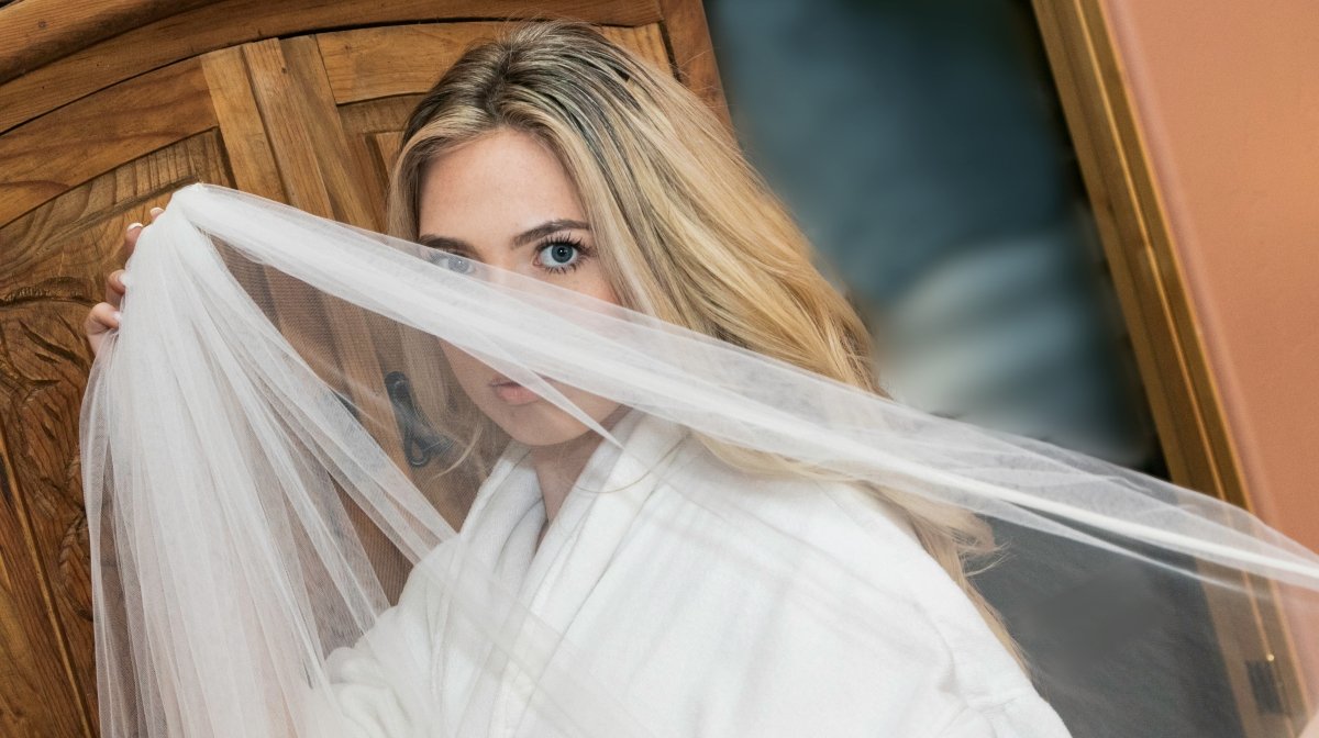 A woman with blonde hair and blue eyes peeks out from behind a sheer white veil posed against a wooden headboard.