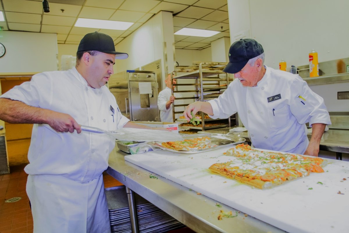 Two chefs in white uniforms and black caps preparing pizza in a commercial kitchen.