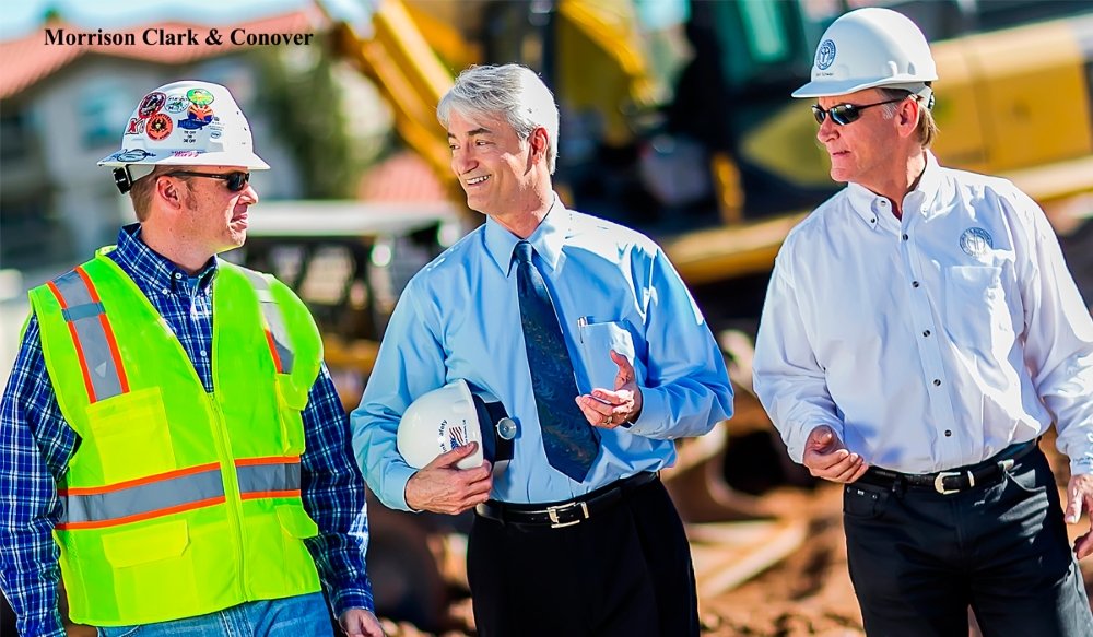 Three men in construction attire and business attire, including hard hats, standing outdoors at a construction site, engaged in conversation.