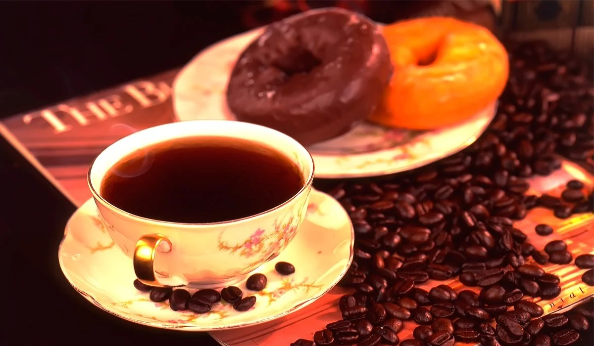 A cup of black coffee in a floral porcelain cup with a gold handle, surrounded by coffee beans, with a plate of two donuts, one chocolate glazed and one plain, in the background.