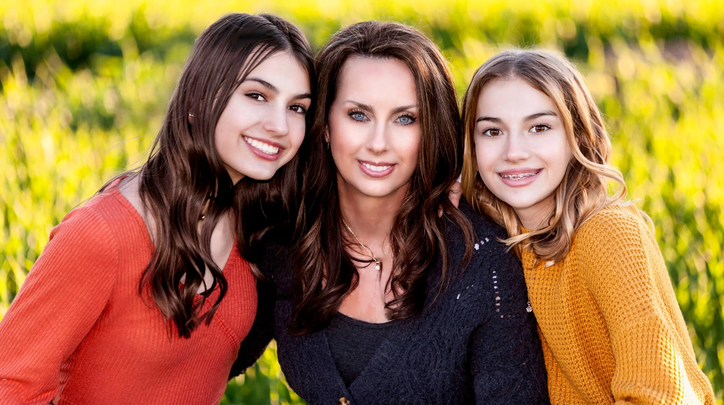 A woman with long brown hair and two young girls, one with long dark hair and the other with light brown hair and braces, smiling together outdoors in front of a green grassy background.