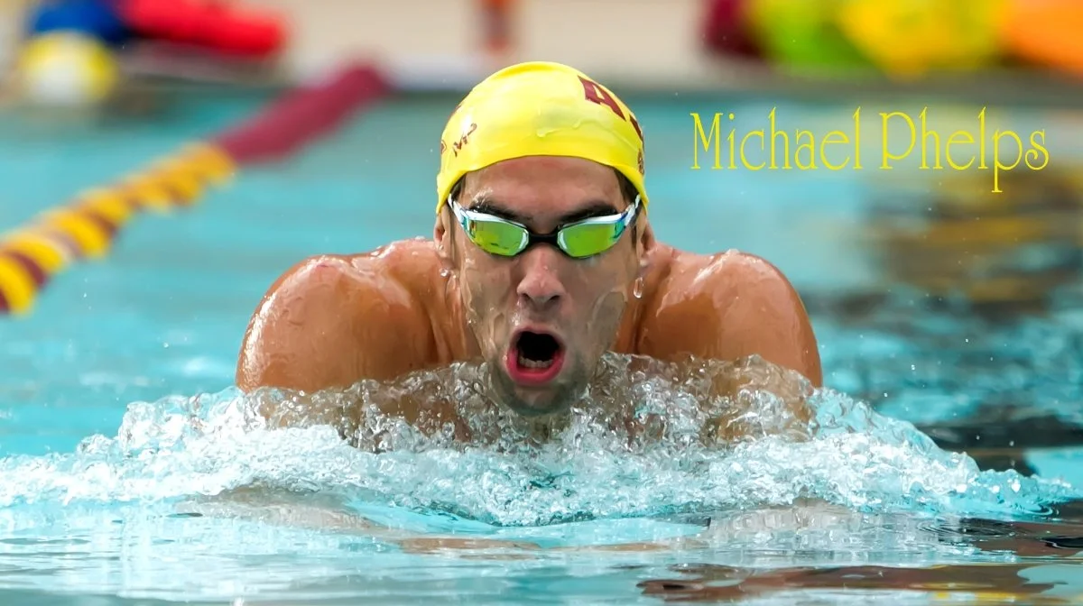 Swimmer wearing a yellow swim cap and goggles, lunging forward in the water during a race, with yellow text saying "Michael Phelps" on the right side.