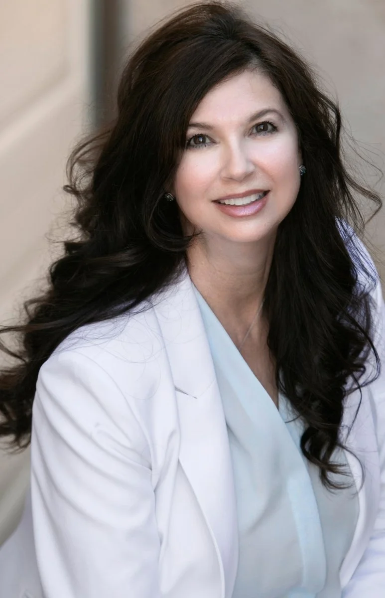 A woman with long dark wavy hair wearing a white medical coat, smiling in a professional portrait.