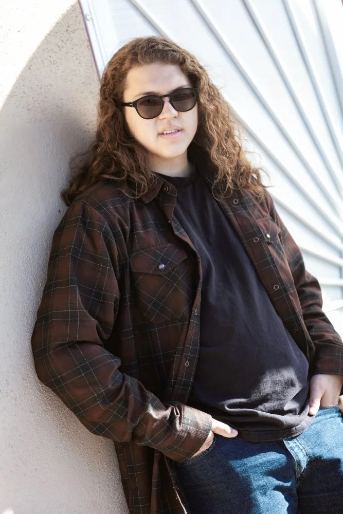 Young woman with curly hair wearing sunglasses, a black shirt, a brown plaid flannel, and blue jeans, leaning against a light-colored wall outside near a building with metallic siding.