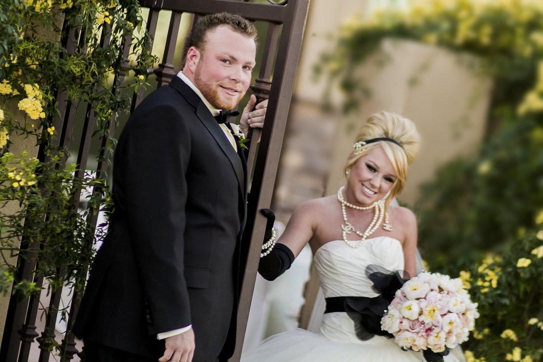 A bride and groom on their wedding day, standing outdoors near a gate with green foliage and yellow flowers in the background. The groom wears a black tuxedo with a bow tie, and the bride wears a white strapless wedding gown with black gloves, pearl 