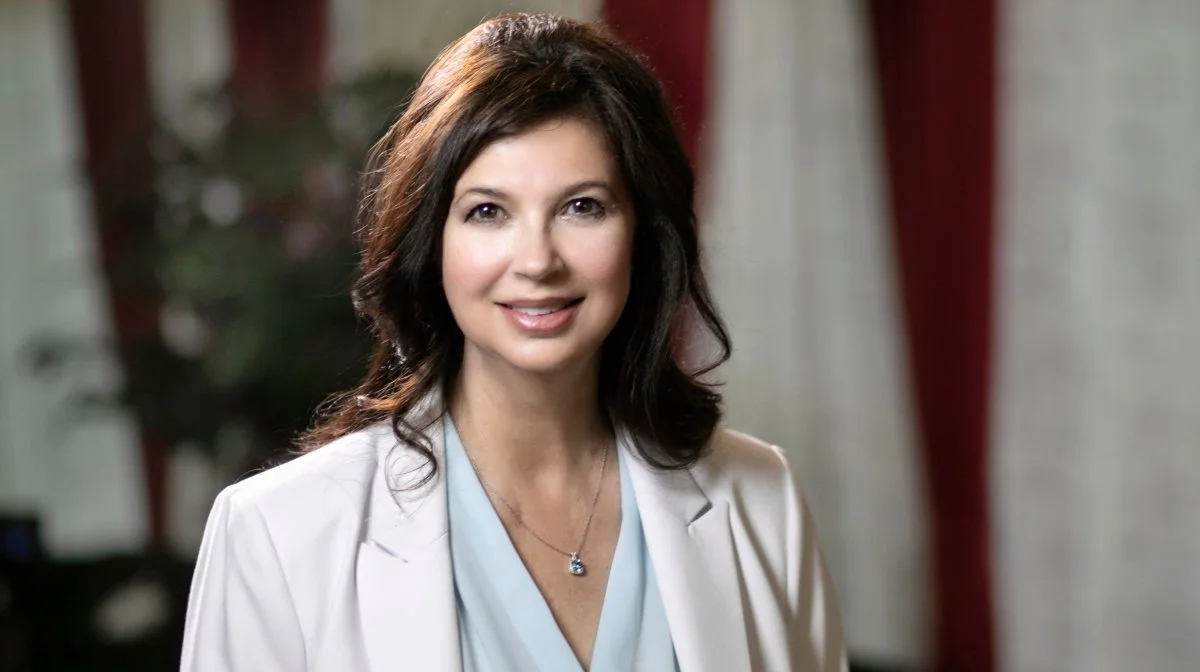 A woman with dark brown hair wearing a white blazer and a light blue top, smiling at the camera, in an indoor setting with blurred background.