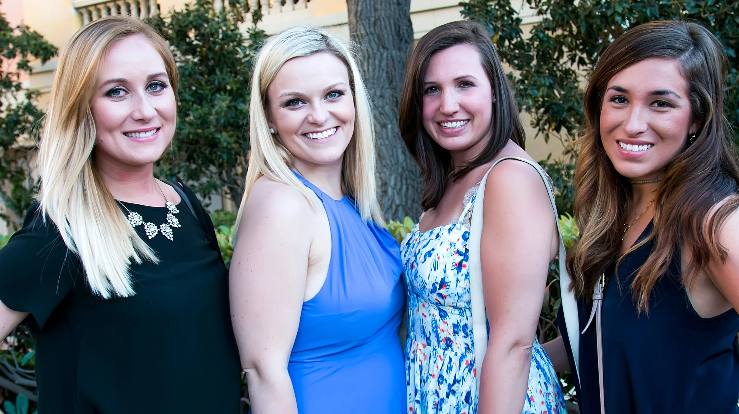 Four young women standing outdoors, smiling, with trees and a building in the background. They are dressed in casual summer clothing.