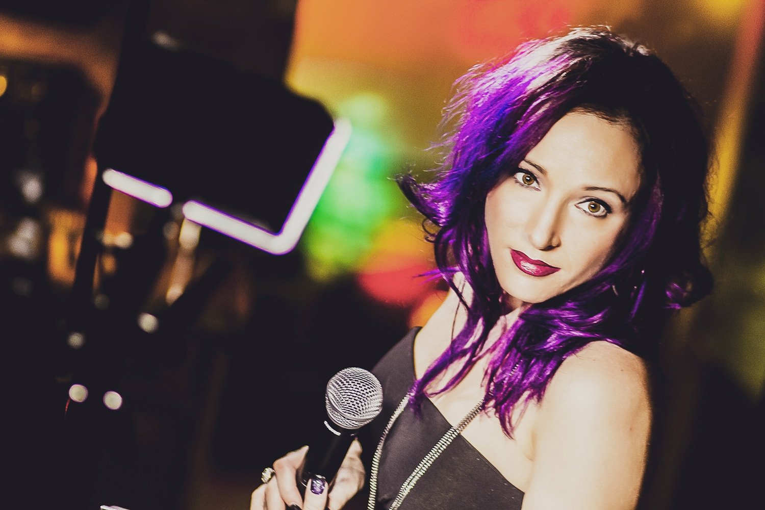 A woman with purple hair holding a microphone, in a dimly lit environment with colorful lights in the background.