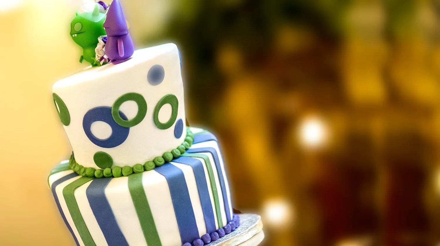Close-up of a colorful birthday cake with a striped pattern in blue, green, and white, topped with a white cube-shaped decoration with circular cutouts, and small purple and green decorations, including two small figurines on top.