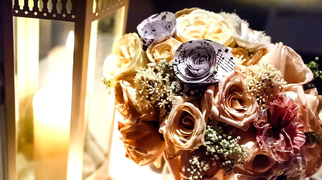 A bouquet of peach and cream-colored roses, small white flowers, and paper roses with musical notes, next to a lit lantern.