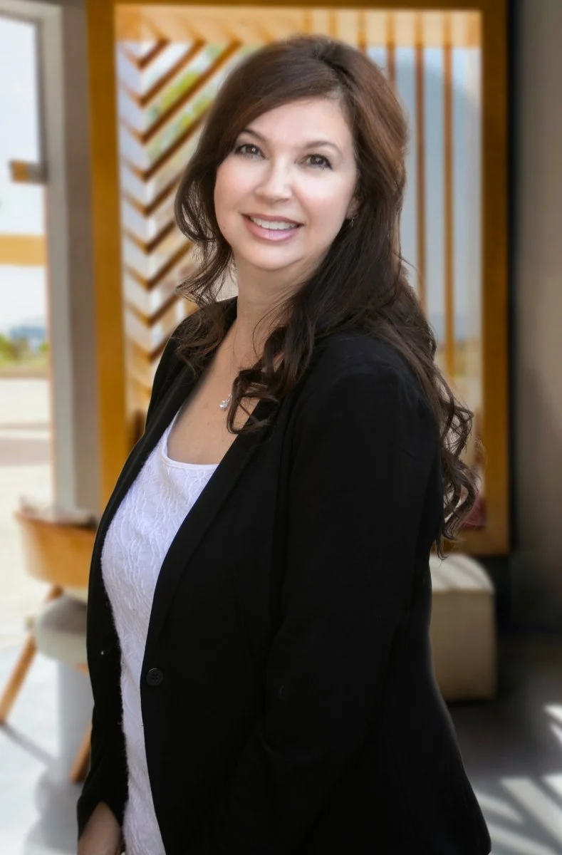 A woman with long dark brown hair wearing a black blazer and white top, smiling in an indoor space with wooden decor in the background.