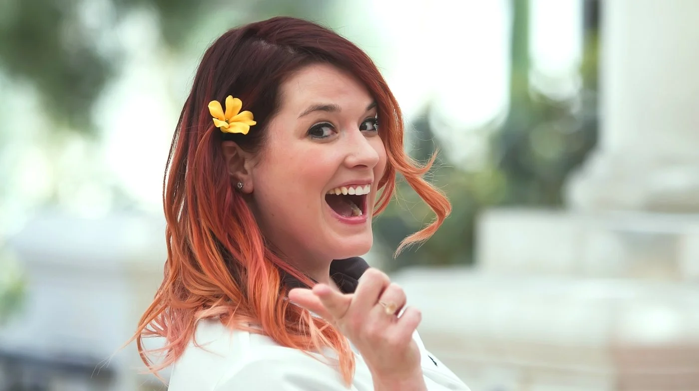 Woman with red hair, yellow flower hair accessory, smiling and pointing at the camera, indoors with blurred background