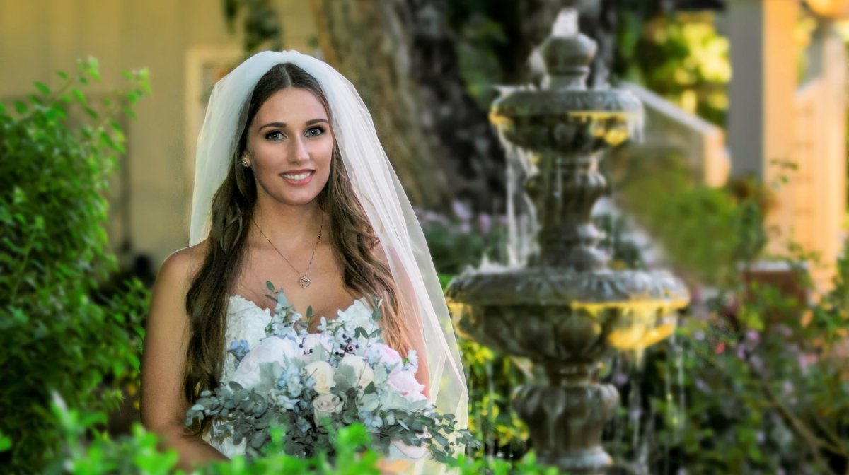 A smiling woman in a wedding dress with a veil holding a bouquet of white and pink flowers, standing outdoors near a fountain and greenery.